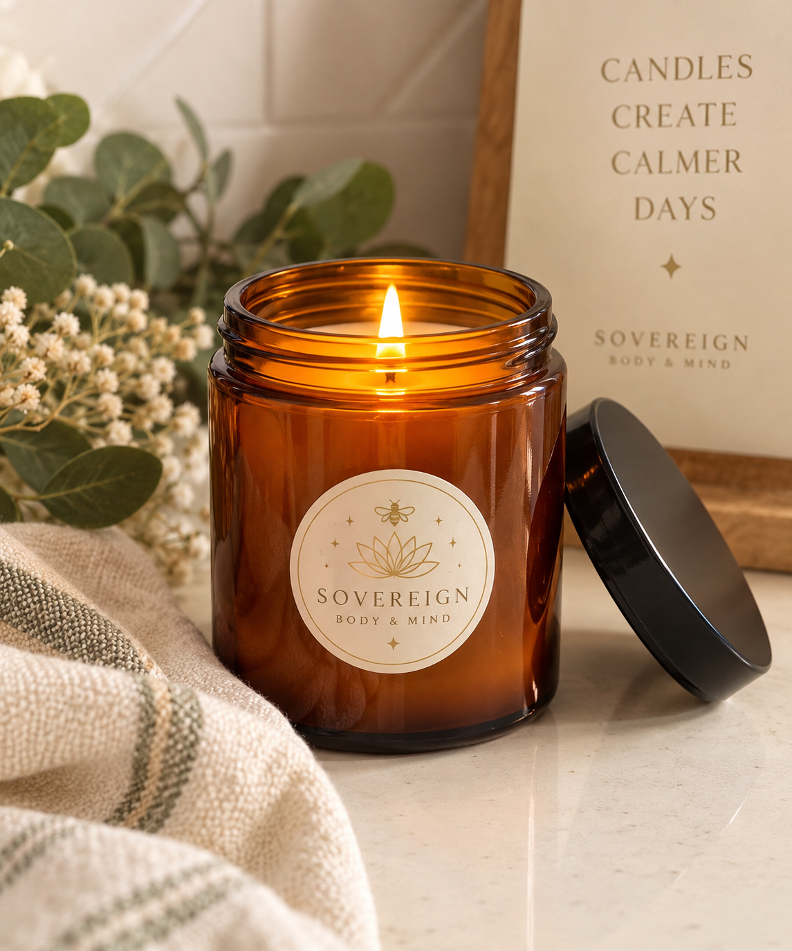 Lit Sovereign Body & Mind amber glass candle beside eucalyptus, dried florals and a linen throw